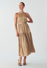 Béžové šaty na krk s čiernymi bodkami, so skladanou sukňou a zaväzovacím pásom. V kombinácii s čiernymi sandálmi na pásky. Hladká textúra látky.