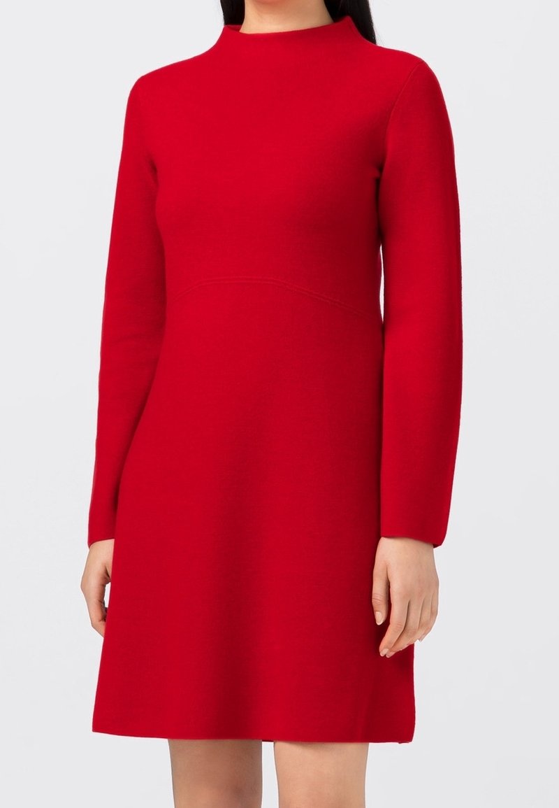 Donna che indossa un abito rosso vivo a maniche lunghe, lungo fino al ginocchio, con uno scollo alto, su uno sfondo grigio chiaro uniforme.