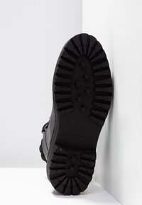 Semelle de botte noire avec des crampons profonds et des motifs en forme d'étoile, positionnée verticalement contre un fond blanc d'angle.