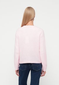 Šviesiai rožinė megzta palaidinė su apvaliu iškirpimu ir ilgomis rankovėmis, turinti švelnią, pūkuotą tekstūrą ir šiek tiek trumpesnį apatinį kraštą.