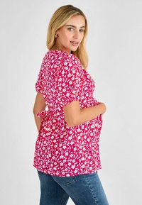 Blusa floreale rosa con maniche a sbuffo legata sul retro, con un motivo di fiori bianchi, abbinata a jeans blu. Tessuto morbido.