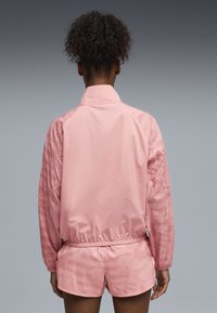 Ροζ αδιάβροχο μπουφάν με ελαστικά μανίκια και ζώνη μέσης. Διαθέτει διακριτικό σχέδιο και ελαφριά, λεία υφή. Εμφανίζεται με ταιριαστό σορτς.