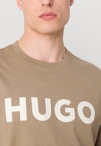 Khaki bavlněné tričko s velkým bílým grafikou "HUGO" na přední straně, s kulatým výstřihem a hladkou texturou.