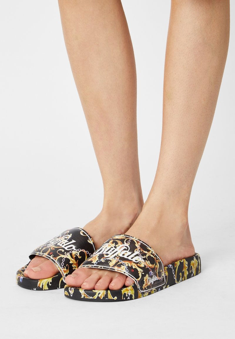 Pieds portant des sandales slides noires avec des motifs ornés dorés et blancs et le mot "Buffalo" sur les brides sur fond blanc.