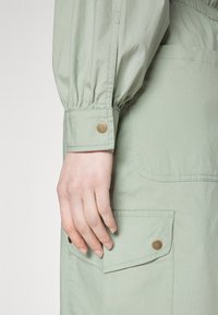 Prenda verde claro con puños de manga fruncidos, que presenta un bolsillo con un botón de presión de bronce. Se puede ver la textura del tejido suave y liso.