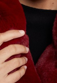 Röd fuskpälskrage med en mjuk textur, kompletterad av en svart stickad tröja under. En hand rör försiktigt vid kragen.