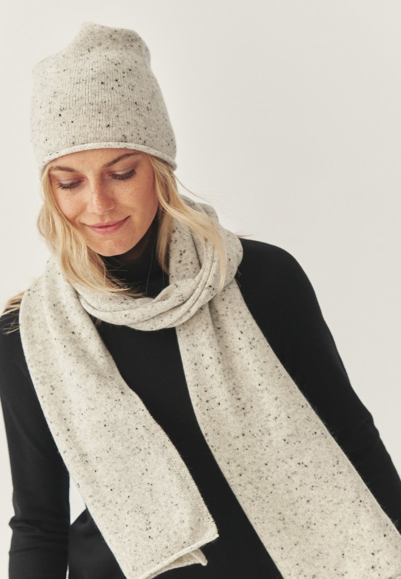 Lysegrå strikket hue med spraglet tekstur kombineret med et matchende tørklæde. Begge er bløde og rektangulære og bæres over en sort top.