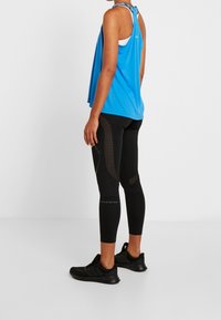 Kvinna i en blå ärmlös topp, svarta mönstrade leggings och svarta träningsskor, stående mot en enfärgad vit bakgrund.