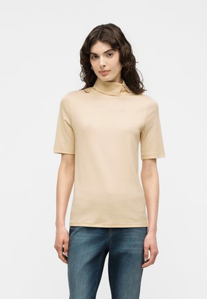 Kvinde med bølget brunt hår iført en beige kortærmet top med høj krave og blå jeans, stående mod en ensfarvet lys baggrund.