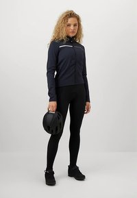 Γυναίκα με σγουρά ξανθά μαλλιά, φορώντας ναυτικό μπλε τζάκετ ποδηλασίας και μαύρες κολάν, κρατώντας ένα μαύρο κράνος ποδηλασίας στο δεξί της χέρι.