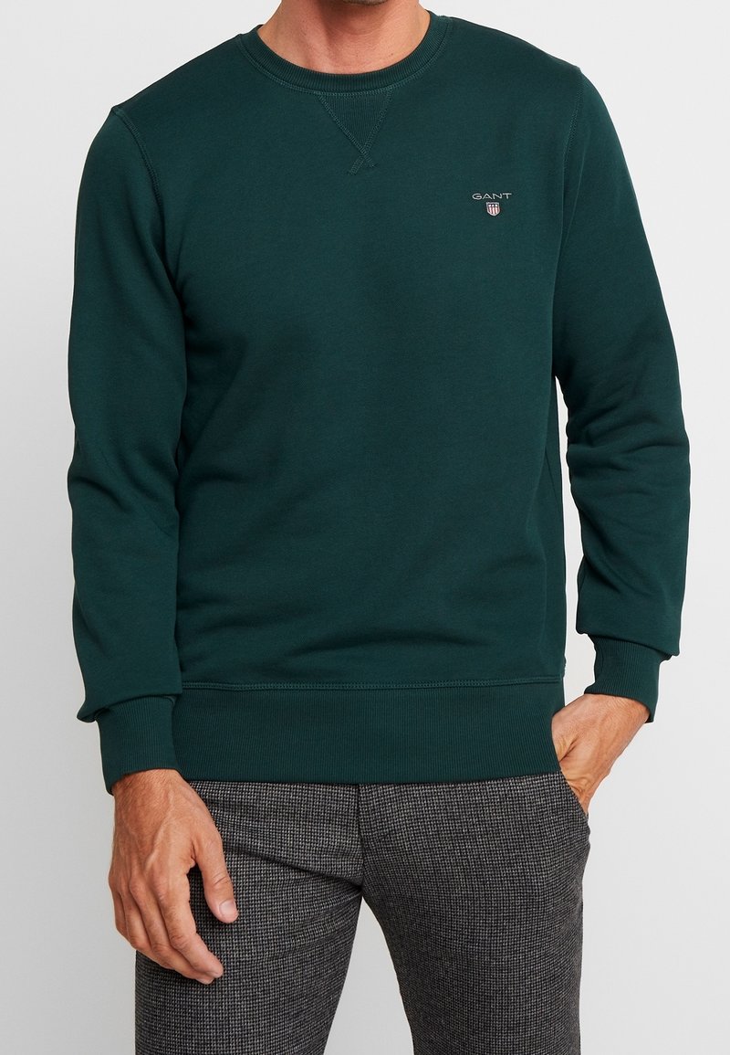 Sweatshirt vert foncé en coton doux, avec col rond, poignets et ourlet côtelés, et un petit logo sur la poitrine.