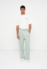 Weißes T-Shirt mit kurzen Ärmeln und Logo, kombiniert mit hellgrünen Jogginghosen mit einem elastischen Bund und Logodetails, getragen mit weißen Schuhen.