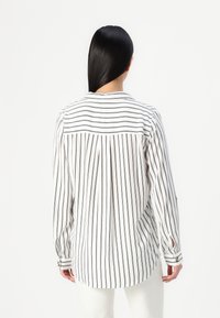 Femme aux longs cheveux foncés portant une chemise blanche à manches longues avec des rayures noires verticales et horizontales, vue de dos.