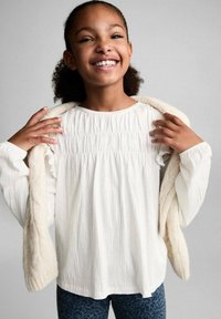 Top bianco e testurizzato con dettaglio arricciato sul colletto, abbinato a un maglione beige chiaro lavorato a maglia drappeggiato sulle spalle e leggings scuri con motivo.