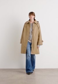 Beige wasserdichter Mantel mit lockerer Passform, Kapuze mit Kordelzug und seitlichen Taschen, getragen über einem grauen Pullover und blauen Schlagjeans.