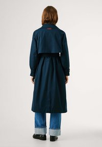 Manteau trench en bleu marine avec une taille ceinturée et un large col, présentant un design court à l'arrière. Associé à un jean bleu et des chaussures noires.