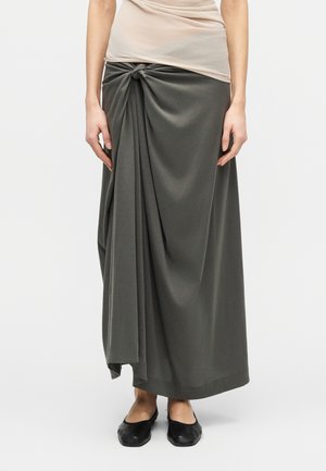 Kvinde iført en lang mørkegrå nederdel med en knude foran og draperet stof, kombineret med sorte flade sko og en beige top.