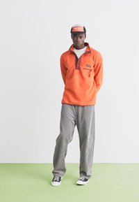 Oranje fleece pullover met een bruine kraag en een borstzak. Gecombineerd met grijs gestreepte broek en zwart-witte sneakers.
