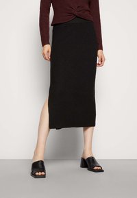 Saia midi preta de malha com uma fenda lateral, combinada com mules de salto aberto pretos. O top é uma camisa de manga longa em bordeaux com textura canelada.