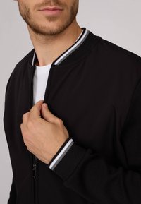 Veste bomber noire avec des poignets côtelés présentant des rayures grises et blanches. Texture de tissu lisse, fermeture éclair à l'avant et design minimaliste.