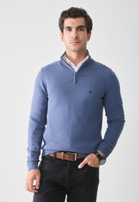 Blå zip-up genser i myk stoff, med en høy krage og ribbestrikkede mansjetter. Kombinert med et brunt skinnbelte og svarte jeans.