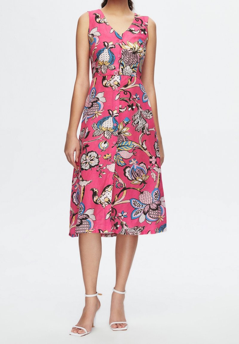 Robe midi rose sans manches avec motif floral et oiseaux coloré, col en V, boutonnée sur le devant, portée avec des talons blancs à bride cheville.
