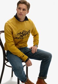 Sudadera color mostaza con gráfico de montaña, combinada con una camisa de cuadros azul marino. Lleva puestos unos jeans azules y botas de cordones marrones.