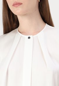 Witte blouse met een hoge hals, met plooien en een kleine zwarte knoopsluiting. Glad, lichtgewicht stof met een subtiele glans.
