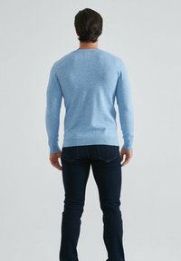 Pull en tricot bleu clair à manches longues, col rond, avec des poignets et un ourlet côtelés. Porté avec un jean foncé, mettant en valeur une silhouette ajustée.