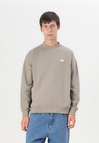 Beige crewneck sweatshirt med ribbestrikkede ermer og kant. Har et lite rødt og hvitt logo-patch på brystet. Båret med blå denimjeans.