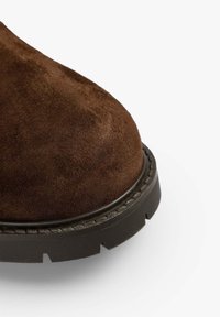 Botas de ante marrón con puntera redondeada y suela gruesa negra con un patrón de relieve texturizado y detalle de costura en contraste.
