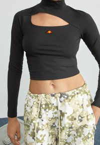 Svart långärmad cropped topp med hög krage och utskärningsdetalj, med en liten färgad logotyp på bröstet. Matchas med mönstrade lösa byxor.