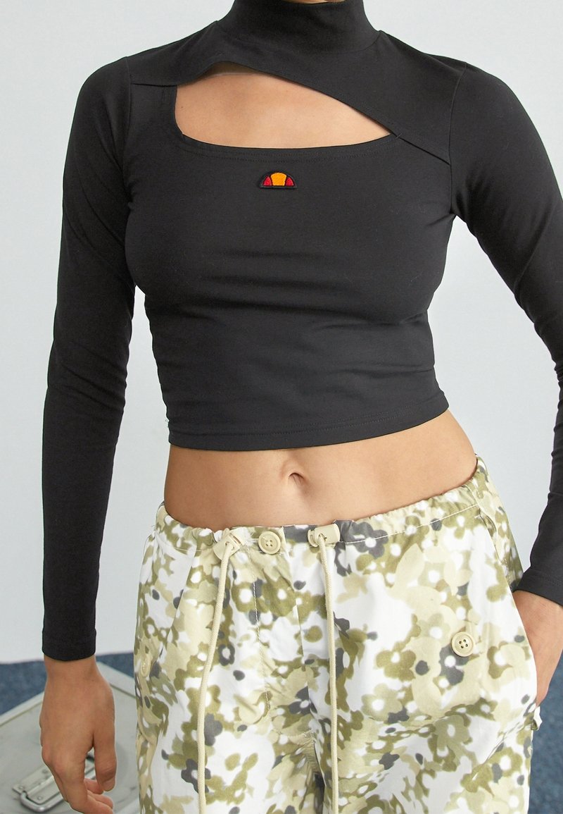 Svart långärmad cropped topp med hög krage och utskärningsdetalj, med en liten färgad logotyp på bröstet. Matchas med mönstrade lösa byxor.