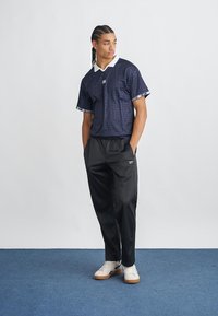Polo din jerseu grafic bleumarin cu guler alb și pantaloni sport negri. Modelul poartă adidași albi cu tălpi bej. Fundal simplu și podea albastră.