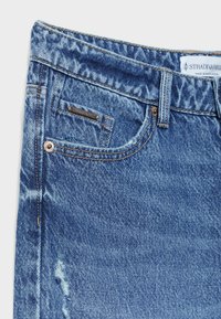 Close-up van de voorzak van een blauwe spijkerbroek met het label "Stradivarius" en metalen klinknagels, waarbij de versleten stoffen details zichtbaar zijn.