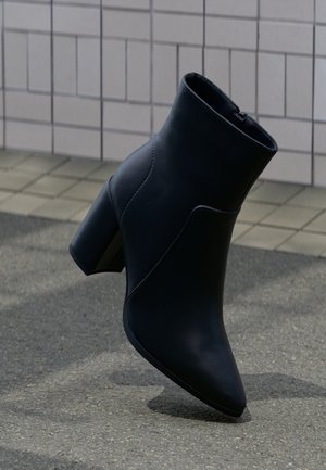 Bottine en cuir noir avec talon bloc, bout pointu et fermeture éclair latérale, posée sur un trottoir rugueux près d'un mur carrelé.