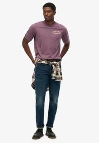 Camiseta de algodón morada con estampado "Superdry Los Ángeles", combinada con jeans de mezclilla azules y una camisa de franela a cuadros atada alrededor de la cintura. Zapatos negros.