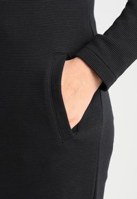 Vêtement en tissu côtelé noir avec une poche latérale. La texture est irrégulière, mettant en valeur des rainures verticales. La coupe semble ajustée et épurée.