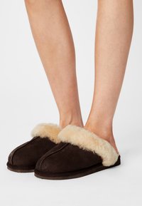 Brune semskede tøfler med en fluffy beige faux fur krave. Designet er åbent foran med flad sål og synlige sømdetaljer.