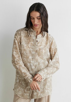 Frau mit langen dunklen Haaren, die ein beige gemustertes, langärmeliges Hemd mit Knöpfen trägt und vor einer schlichten weißen Wand die Manschette mit den Händen anpasst.