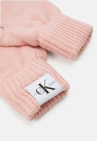 Strikkede fingerløse hansker i myk rosa, med ribbestrikkede håndleddsmanchetter og en hvit etikett med svart tekst som sier "Calvin Klein Jeans."