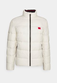 Weißer gesteppten Pufferjacke mit schwarzem Reißverschluss und Taschen, Stehkragen und kleinem roten quadratischen Etikett auf der linken Brust.