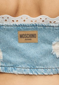 Κοντινό πλάνο σε τζιν Moschino από τζιν με φθαρμένη λεπτομέρεια και λευκή δαντέλα με κυματιστό μοτίβο στη μέση επάνω σε γυμνό δέρμα.