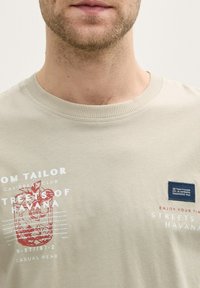 Camiseta de algodón color beige claro con cuello redondo, que presenta un diseño gráfico rojo y texto blanco. Incluye una etiqueta de tela azul en el pecho.