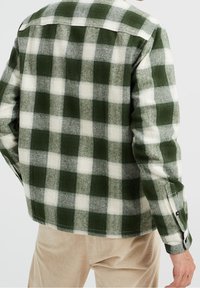 Man wearing green and white checkered long-sleeve shirt and beige corduroy pants, shown from the back with right arm relaxed.