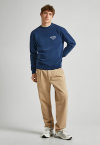 Tumši zila džemperis ar apaļu kakla izgriezumu, kuru papildina logo uzšuvums, apvienots ar beigu krāsas brīvā stila biksēm un baltām treniņbiksēm ar pelēkiem akcentiem.