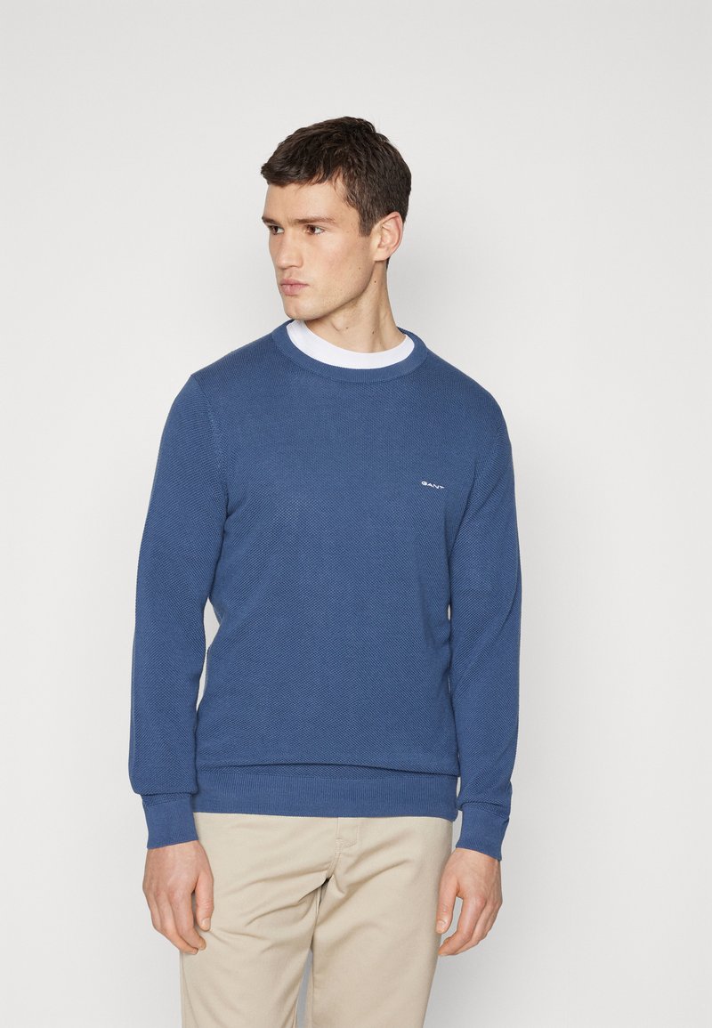 Joven vestido con un suéter azul de punto con cuello redondo sobre una camisa blanca, combinado con pantalones beige, de pie frente a un fondo blanco liso.