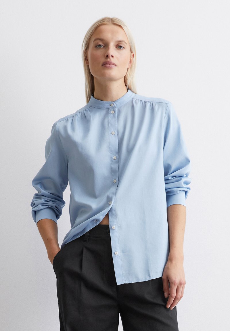 Light blue, long-sleeve button-up shirt with a stand collar, relaxed fit, and gathered detailing, paired with black tailored pants.