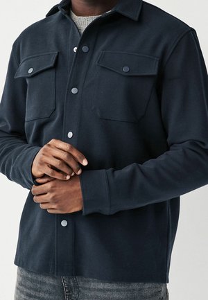 Mann trägt eine dunkelblaue Jacke mit Knöpfen und zwei Patten-Brusttaschen über einem grauen Pullover und richtet mit beiden Händen eine Manschette.