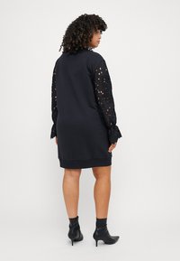 Abito nero con maniche lunghe adornate in pizzo e una silhouette semplice. Il tessuto appare setoso con un orlo sotto il ginocchio. Indossato con stivali neri alla caviglia.